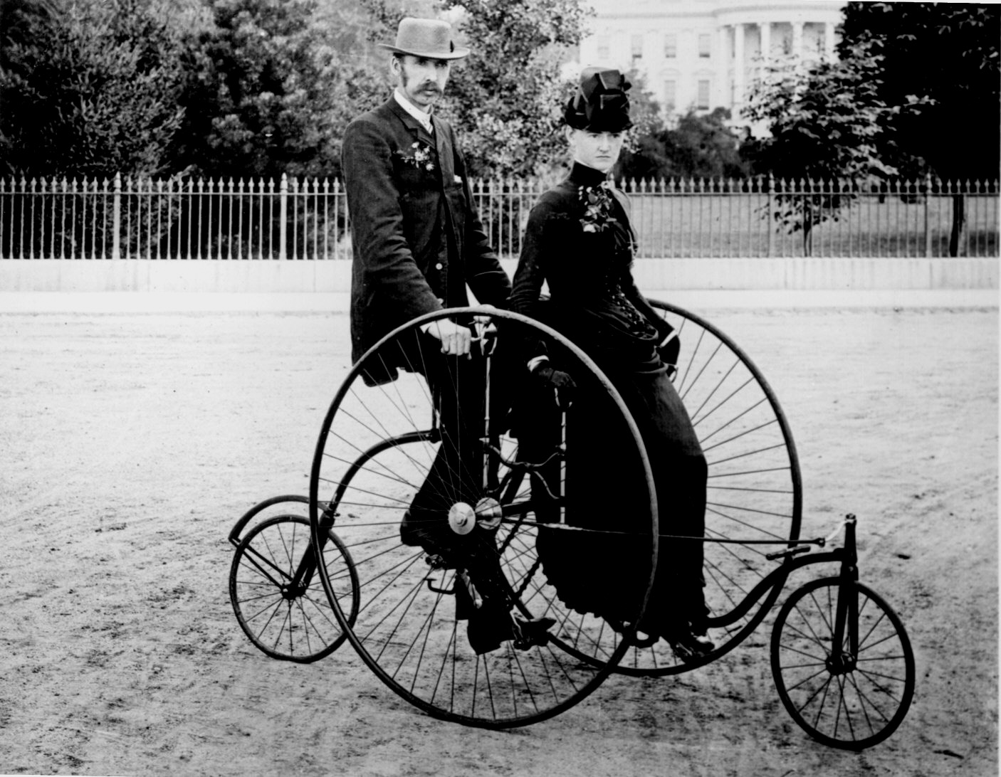 Ein historisches Schwarz-Weiß-Foto aus dem 19. Jahrhundert zeigt ein elegant gekleidetes Paar auf einem ungewöhnlichen Hochrad-Tandemfahrrad. Der Mann trägt einen Anzug mit Hut, die Frau ein langes, dunkles Kleid mit Hut und sitzt seitlich auf dem Beifahrersitz. Beide sitzen zwischen zwei großen Vorderrädern und kleineren Hinterrädern auf einer staubigen Straße, vermutlich vor dem Weißen Haus in Washington D.C., das im Hintergrund hinter einem Eisenzaun sichtbar ist. Das Fahrzeug wirkt wie ein frühes Beispiel mechanischer Mobilität.