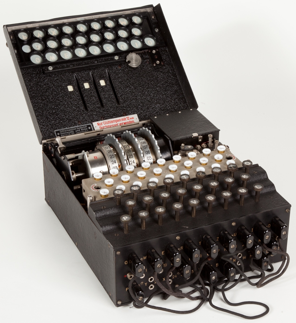 Ein historisches Foto der deutschen Enigma-Verschlüsselungsmaschine aus dem Zweiten Weltkrieg. Die Maschine besteht aus einer Tastatur mit mehreren Reihen schwarzer Tasten, darüber befinden sich drei sichtbare Walzen (Rotoren) mit Zahlen von 01 bis 26. Im oberen Bereich ist ein Leuchtfeld mit runden Lampen zu sehen, die zur Anzeige der verschlüsselten Buchstaben dienten. Darunter erkennt man das sogenannte Steckerbrett (Plugboard) mit mehreren Kabeln zur manuellen Verbindung von Buchstabenpaaren. Das Gehäuse ist schwarz und metallverstärkt, die Bedienung erfolgte mechanisch-elektrisch.