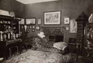Schwarz-Weiß-Fotografie eines opulent eingerichteten Zimmers mit orientalischem Teppichboden, zahlreichen Antiquitäten und Kunstobjekten. Im Zentrum steht eine reich gemusterte Couch mit Decken und Kissen, davor ein kleiner Hocker. Die Couch ist von dekorativen Teppichen umrahmt, die auch an der Wand hängen. Umgeben ist der Raum von voll bestückten Regalen, einem Tisch mit Figuren und Blumen sowie zahlreichen Bildern und Fotografien an den Wänden. Es handelt sich um Freuds ikonisches Behandlungszimmer in Wien, aufgenommen 1938 von Edmund Engelman.