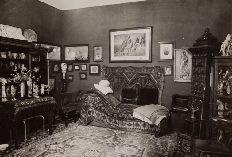 Schwarz-Weiß-Fotografie eines opulent eingerichteten Zimmers mit orientalischem Teppichboden, zahlreichen Antiquitäten und Kunstobjekten. Im Zentrum steht eine reich gemusterte Couch mit Decken und Kissen, davor ein kleiner Hocker. Die Couch ist von dekorativen Teppichen umrahmt, die auch an der Wand hängen. Umgeben ist der Raum von voll bestückten Regalen, einem Tisch mit Figuren und Blumen sowie zahlreichen Bildern und Fotografien an den Wänden. Es handelt sich um Freuds ikonisches Behandlungszimmer in Wien, aufgenommen 1938 von Edmund Engelman.
